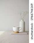Small photo of Cup of coffee and vase with gypsophila flowers on table with linen tablecloth. White wall on background. Still life nordic, Scandinavian home decor
