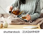 Woman dressed in knitted sweater pours hot tea from glass teapot into mug while sitting in bed. Morning breakfast in cozy home bedroom interior. Hygge, warm, autumn concept.