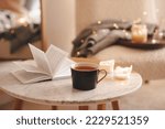 Small photo of Cup of tea with paper open book and burning scented candles on marble table over cozy chair and glowing lights in bedroom closeup. Winter holiday season.