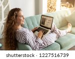 Portrait of beautiful smiling girl doing shopping on internet at home. Cheerful lady laying on sofa in pretty pyjamas. Morning and technology concept. Blurred background