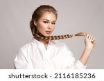 Glamourous young blond caucasian girl with healthy shiny long hairs in pigtail hold braid in hand. Alluring female with stylish plait posing on gray studio background. Haircare treatment cosmetics ad