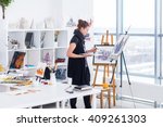 Female painter drawing in art studio using easel. Portrait of a young woman painting with aquarelle paints on white canvas, side view portrait
