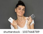 Portrait of pretty doubt young female can't choose modern cellphone, making insure gestures with two smart phones and looking up, isolated over gray background. copy space fot your text or advertising