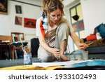Small photo of Horizontal image of a pretty female artist sitting on the floor in the art studio and painting on paper with a brush. A woman painter with glasses painting with watercolors in the workshop.