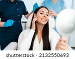 Small photo of Beautiful and happy young woman sitting in medical chair and looking in the mirror. She is satisfied after successful beauty treatment with hyaluronic acid fillers or botulinum toxin injections.