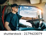 Small photo of Handsome experienced male truck driver with a hat sitting and driving his truck. Professional transportation and truck drivers concept. Side view.