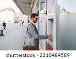 Small photo of Handsome middle age businessman with eyeglasses standing on city street and using ATM machine to withdraw money from credit or debit card.