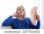 Small photo of A woman in her fifties sitinds fanning herself with a handheld fan, her face flush with the heat of menopause. Mature woman experiencing hot flush from menopause at home