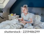 Young excited woman in home clothes passionately watching Tv or movie with bated breath. Girl eating ice cream, holding tissue while sitting on bed at home alone. Leisure, relaxation time