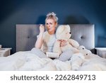 Worried, upset woman hugging big teddy bear, eating ice cream while watching TV in bed. Sad lady over breakup or relationship problems, feeling depressed and lonely. Jamming of negative emotions.
