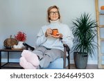 Middle aged woman relaxing with cup of hot drink in scandy style hygge interior home with fall mood interior decor. Lady dreaming, enjoy calm mood without stress, wellbeing alone. Cozy autumn at home.