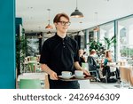 Small photo of Portrait of young smiling affable waiter carrying on tray with coffee cups to client table in cafe. Hospitality service job. Occupation and work concept. Part-time work for students. Selective focus.