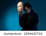 Hiding behind a mask, a young woman in a dark hoodie hides her face with a mask, self-identification problems and impostor syndrome. Portrait in the studio on a dark gray background.