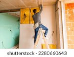 Worker on wooden ladder holds professional manual power tool ACU drill and tightens the screws on plasterboards installed on the partition dry wall insulated with mineral wool.
